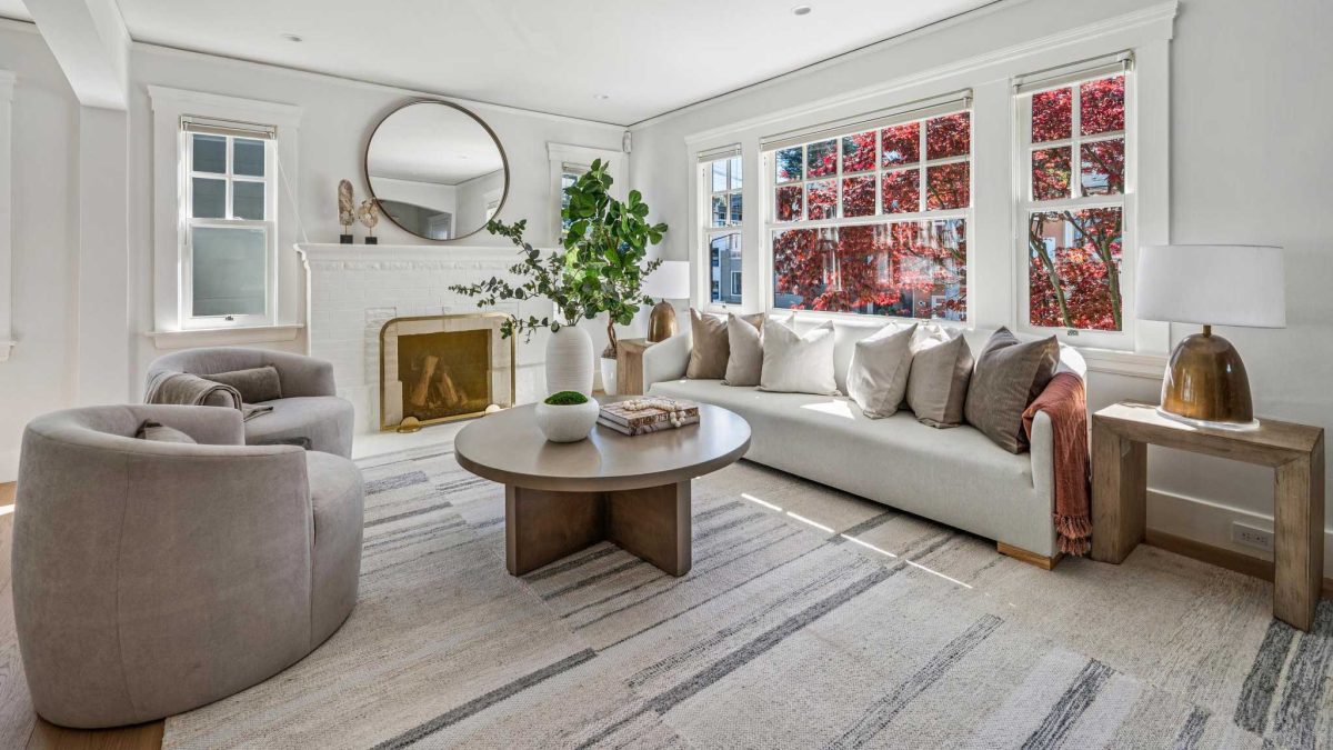 Luxurious living room with modern furniture, neutral accents, fireplace, mirror above mantle, and red leafed tree outside of a picture window